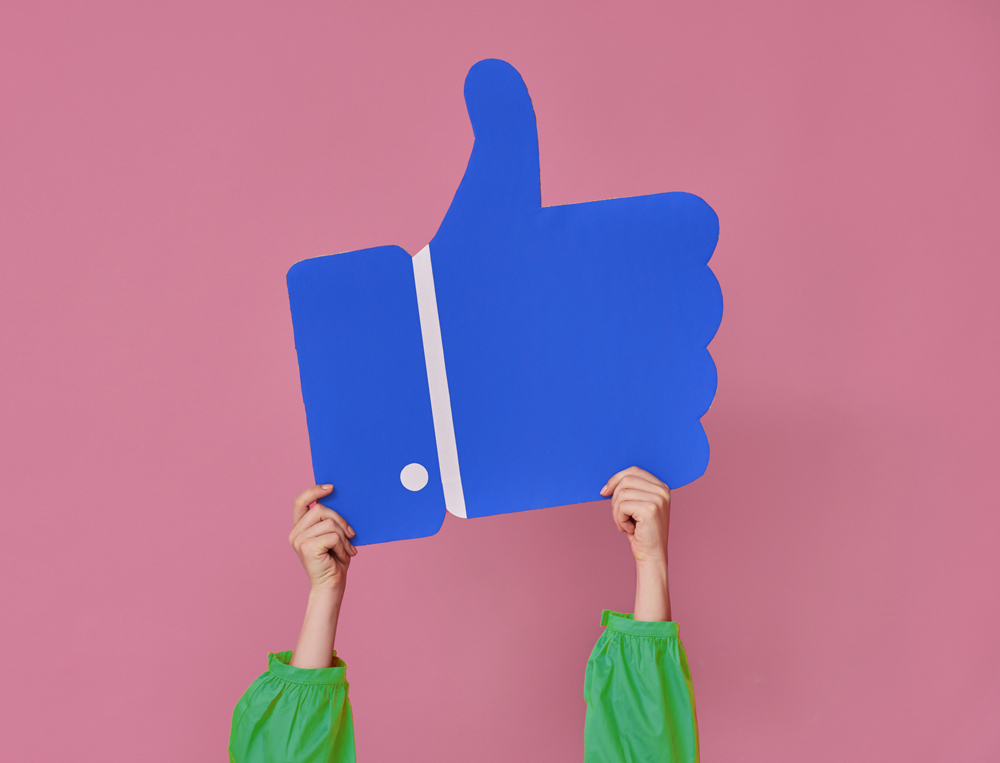 Pink background with two hands holding a cartoon cutout of a thumbs up social media 'like'