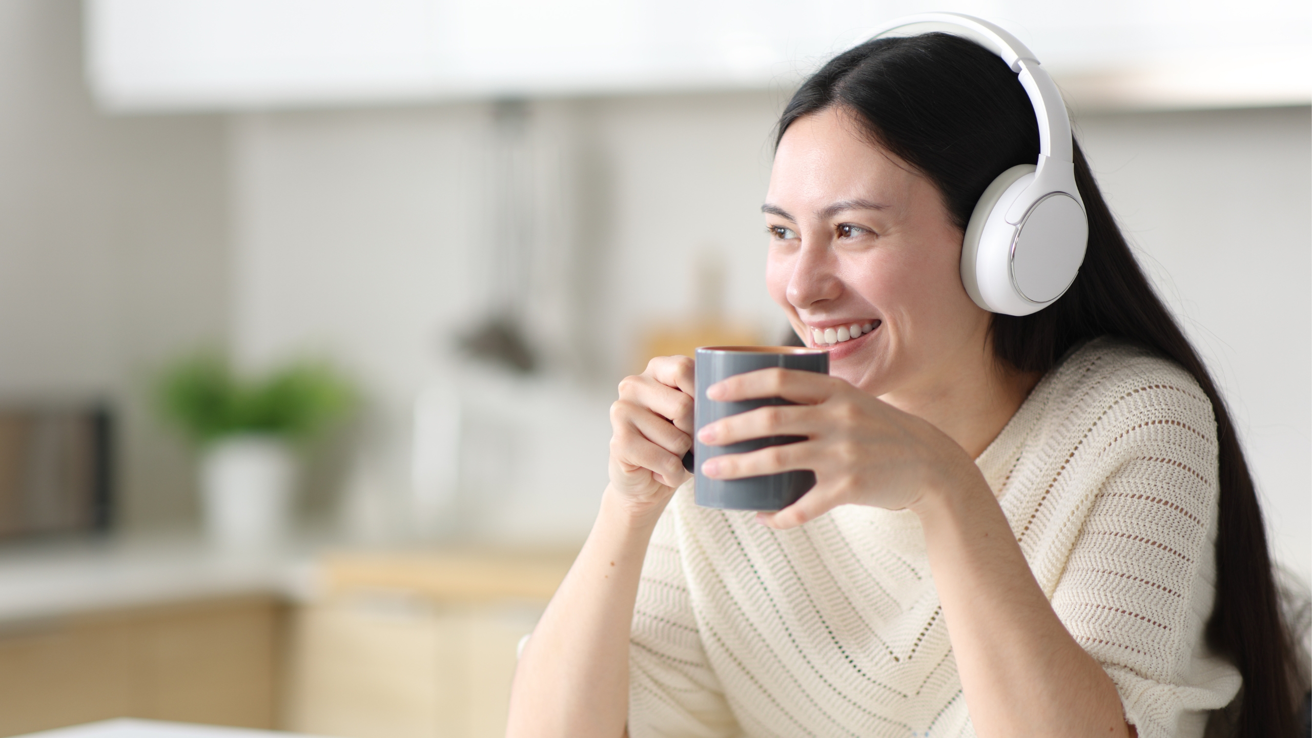 A woman wearing white headphones smiles while holding a mug with both hands, sitting in a bright kitchen. She appears relaxed, enjoying a drink and listening to a podcast about how to set up engagement ads on Facebook.