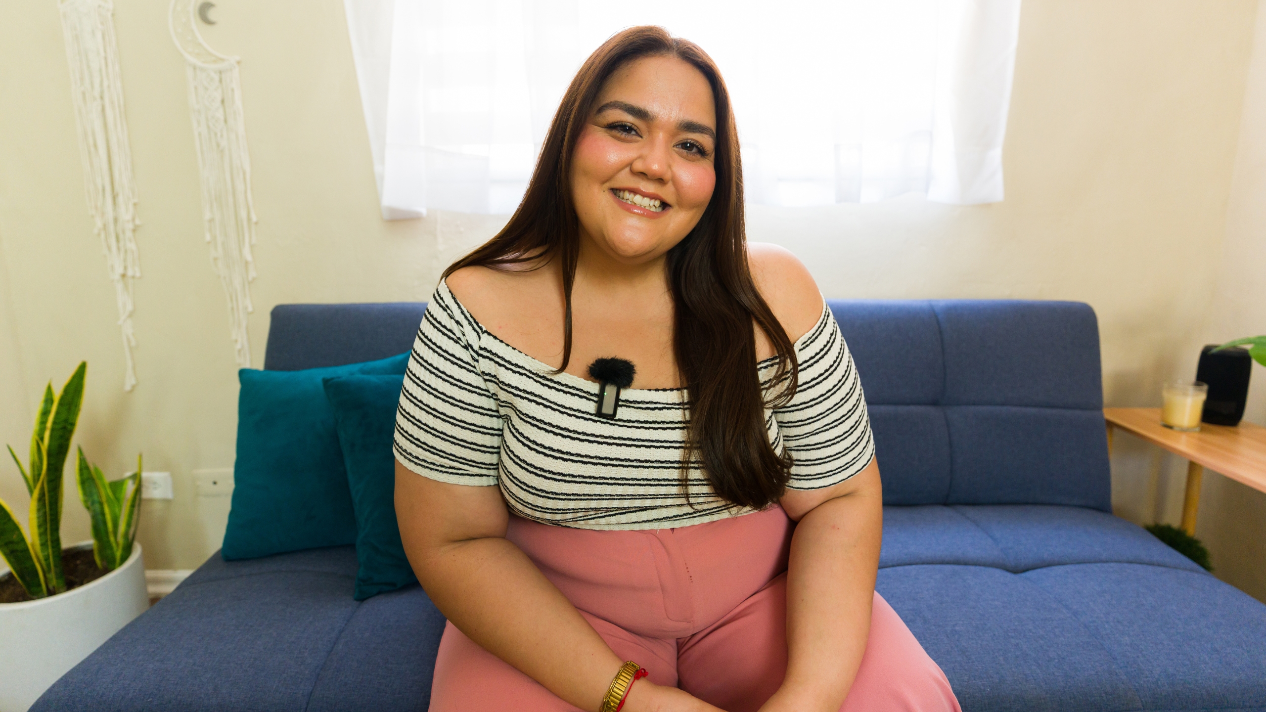 A smiling woman with long brown hair, wearing a striped off-shoulder top and pink pants, sits on a blue sofa in a bright room with plants and a window—perfect inspiration for pretty ads that convert.