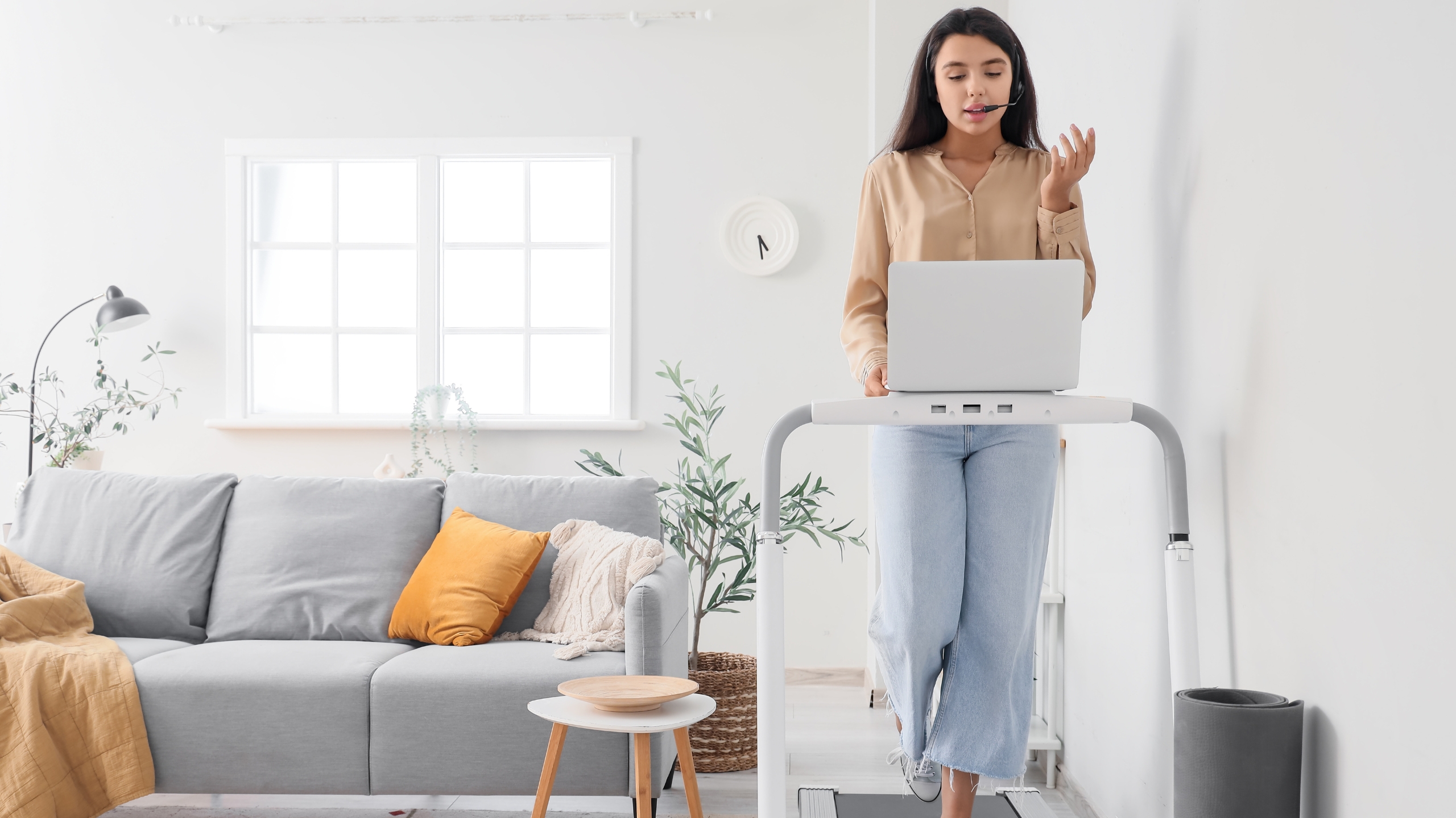 A woman works on a laptop placed on a treadmill desk in a bright living room with a gray sofa, yellow pillow, small round table, and plants. She appears to be talking, possibly discussing $1 leads or lead generation on a video call.