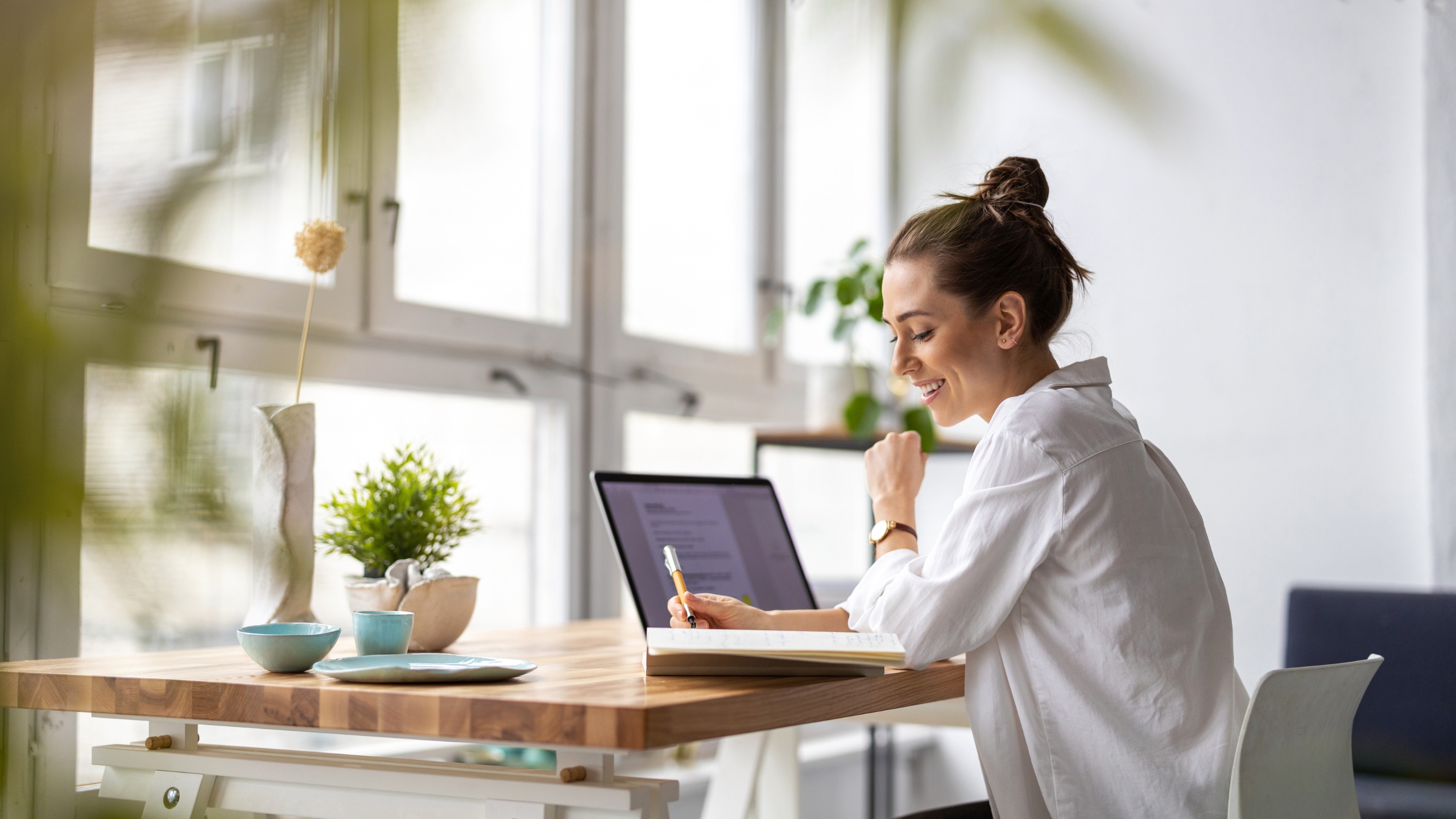 A woman sits at a wooden desk by a window, smiling as she writes in a notebook with a laptop open in front of her, enjoying the predictability of her bright, plant-filled space decorated with simple pottery.