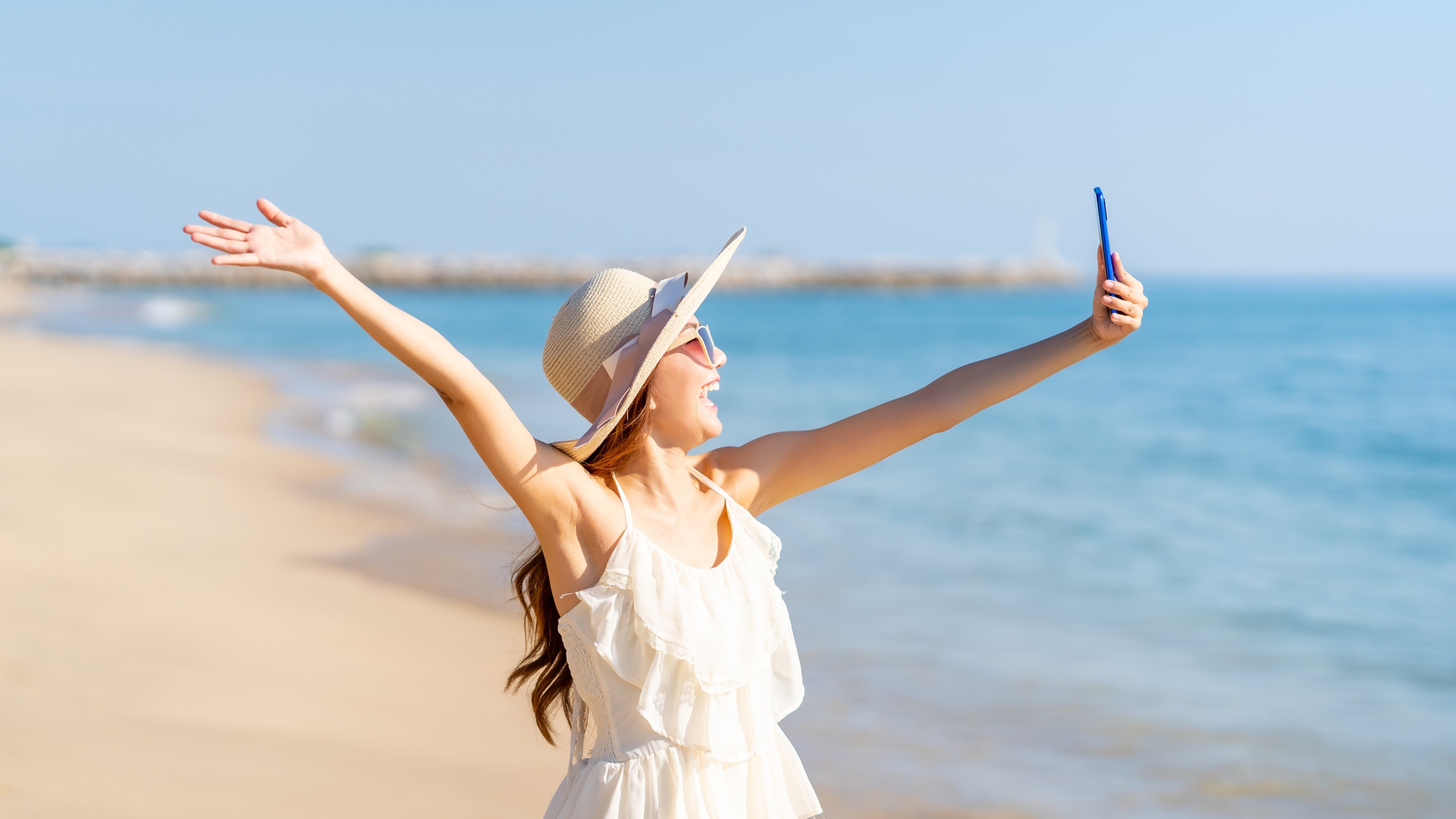 A woman in a white dress and sunhat stands on a sandy beach, smiling and holding up her phone for a selfie—embracing tech with confidence as she raises her other arm joyfully. The blue sea and sky are in the background.