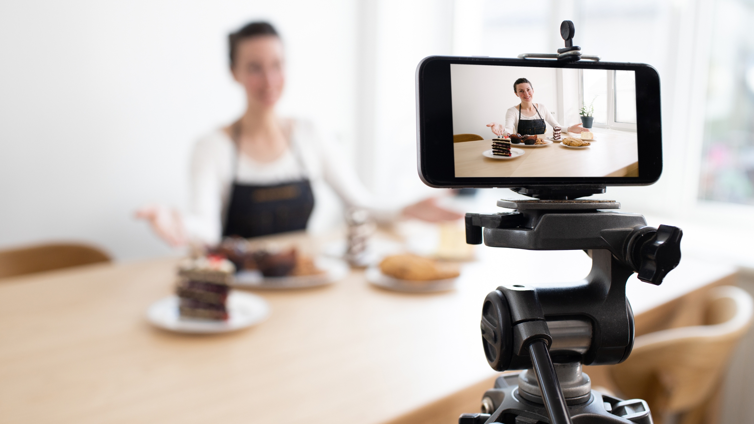 A person wearing an apron sits at a table with desserts, filming themselves with a smartphone on a tripod, likely recording a baking or food vlog in a bright, minimalist room to showcase creative marketing techniques.