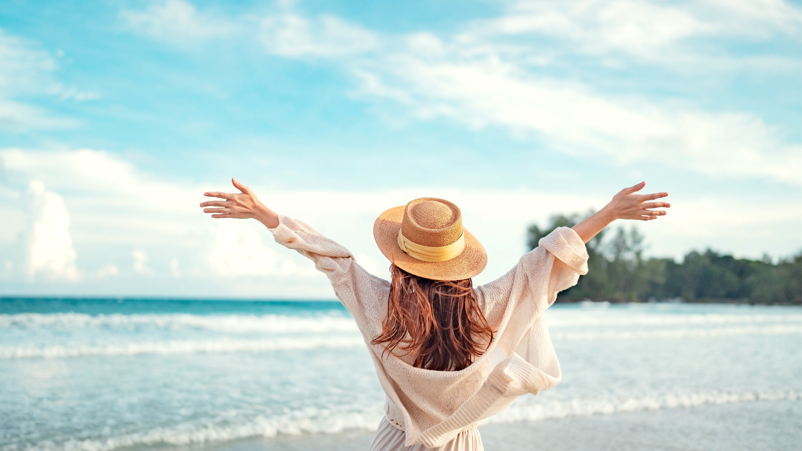 A woman wearing a hat and light clothing stands on a beach with her arms outstretched, facing the ocean—free as the open sky, like reaching for $91K in sales after a successful list-building ad. Trees line the shoreline in the background.