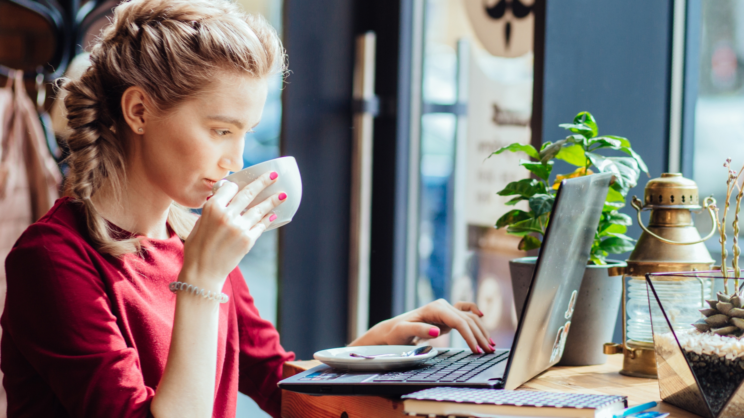 A woman with braided blonde hair, wearing a red top, sits at a wooden table in a café, sipping from a white cup and researching how to set up engagement ads on Facebook while working on her laptop. A small plant and brass lantern are beside her.