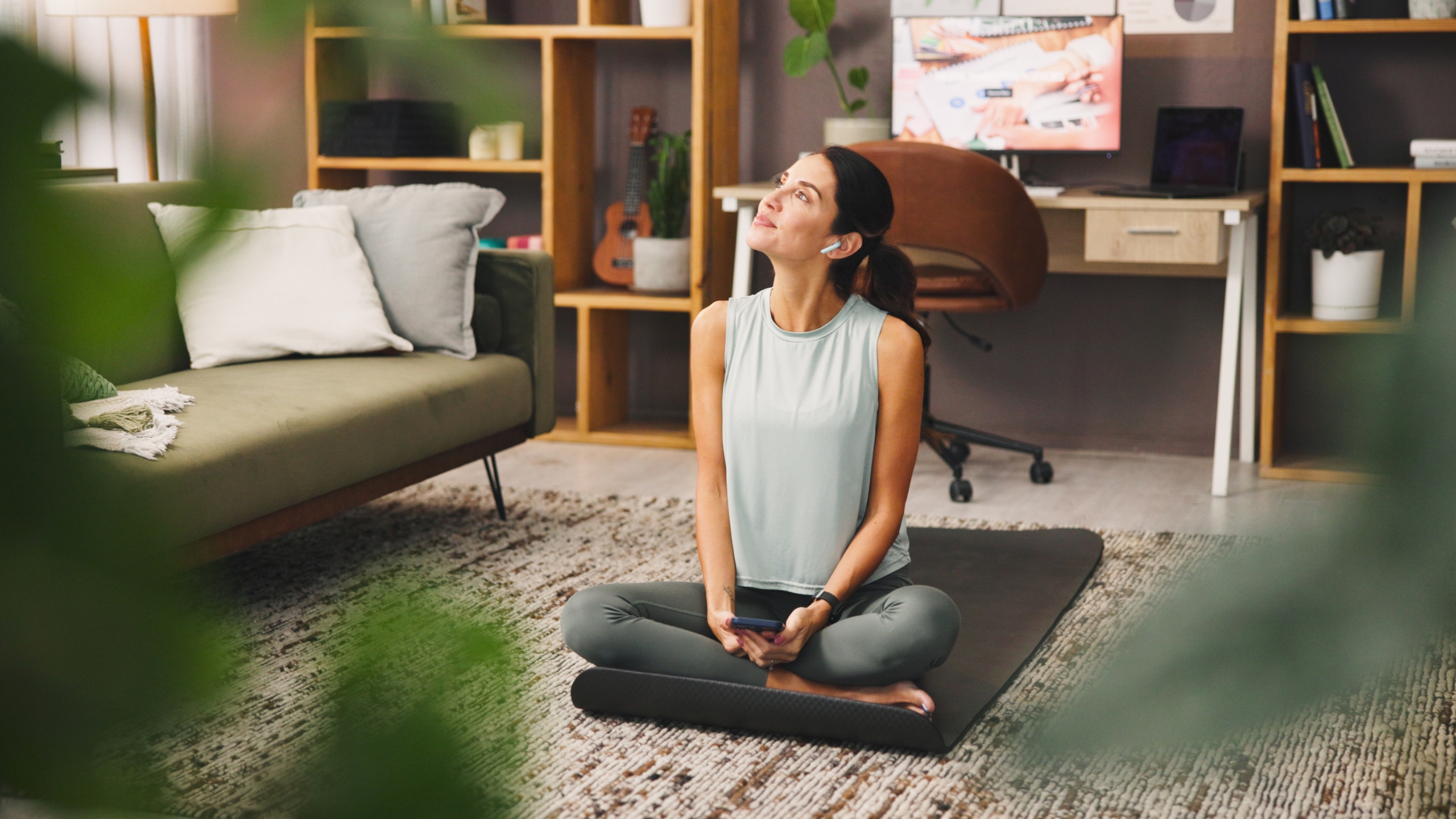 A woman sits cross-legged on a yoga mat in a cozy living room, holding a phone and looking up with a relaxed expression—perhaps reviewing a Lead Magnet Audit. She wears workout clothes and wireless earbuds, with a desk and shelves in the background.