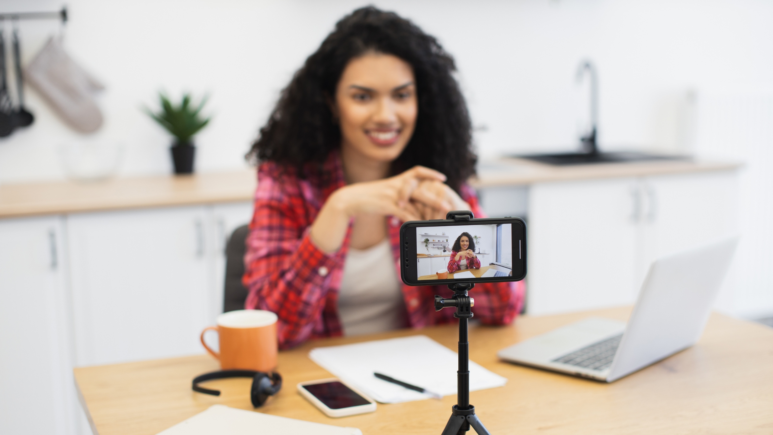 A woman with curly hair sits at a table with a laptop, phone, notebook, and orange mug, recording herself using a smartphone on a tripod in a bright kitchen setting while discussing Black Friday discounting.
