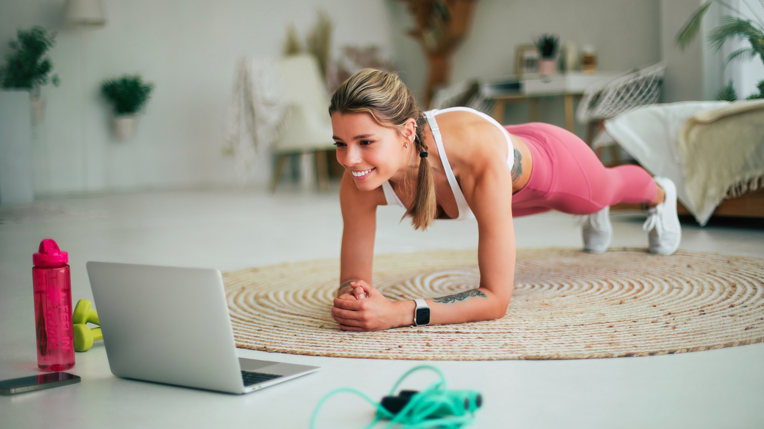 A woman in athletic wear smiles while holding a plank position on a round rug, looking at a laptop in a bright room—perhaps researching how to fix feast or famine revenue cycles amid gym equipment and lush plants.