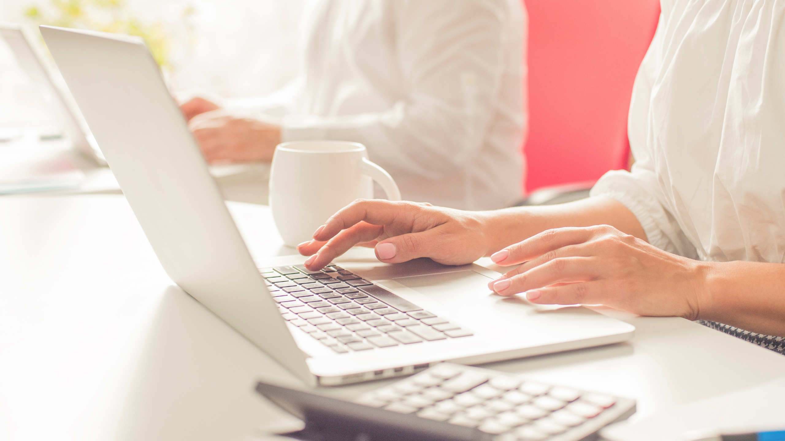 A person types on a laptop at a bright desk with a coffee mug and calculator nearby, planning Black Friday strategies. Another person is blurred in the background, also working at a computer. The scene is well-lit and professional.
