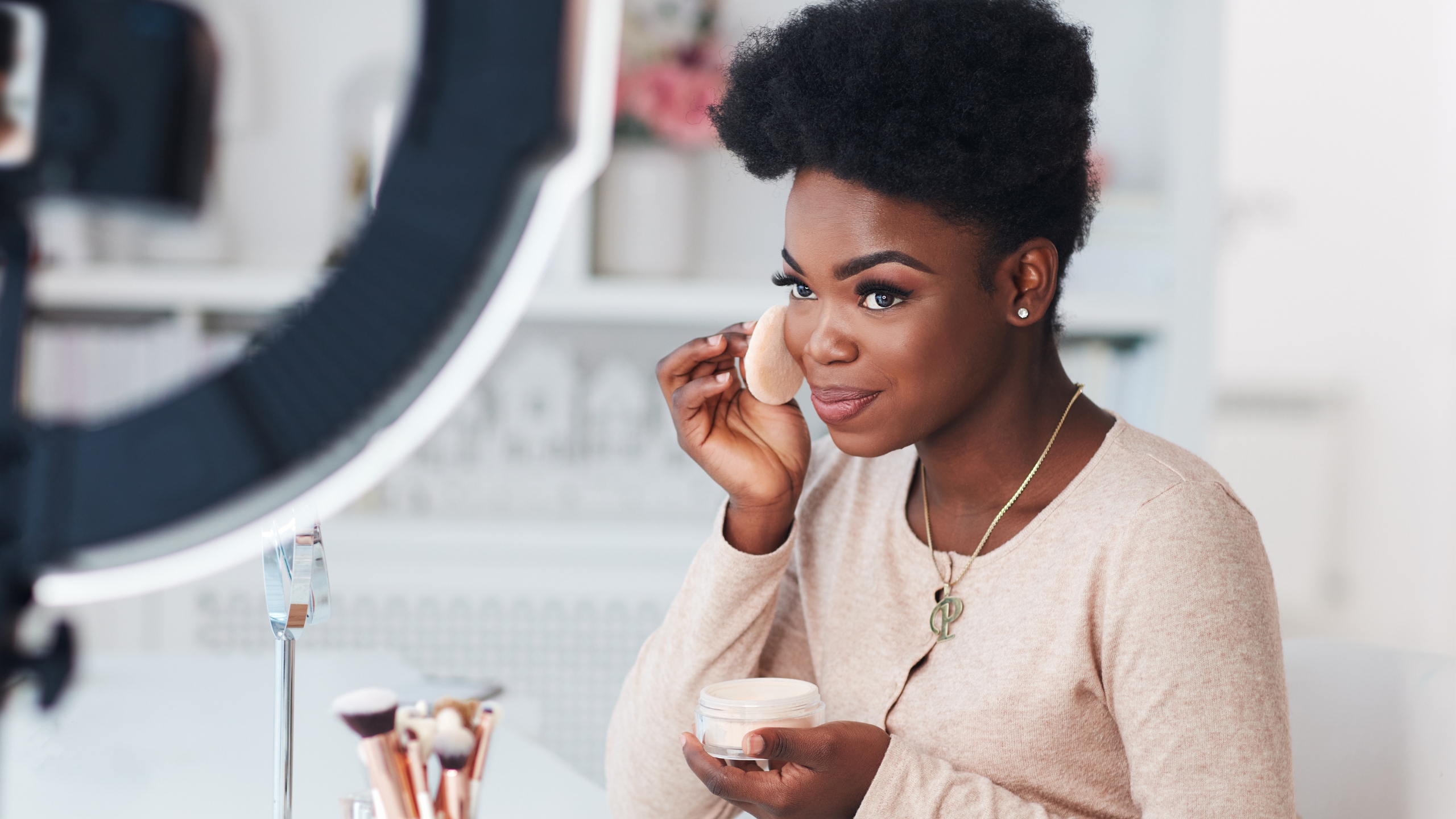 A woman sits in front of a ring light and applies makeup with a powder puff, holding a container of powder. Makeup brushes are on the table nearby. Focused and stylish, she explores proven income streams 2026 to make money between course launches.
