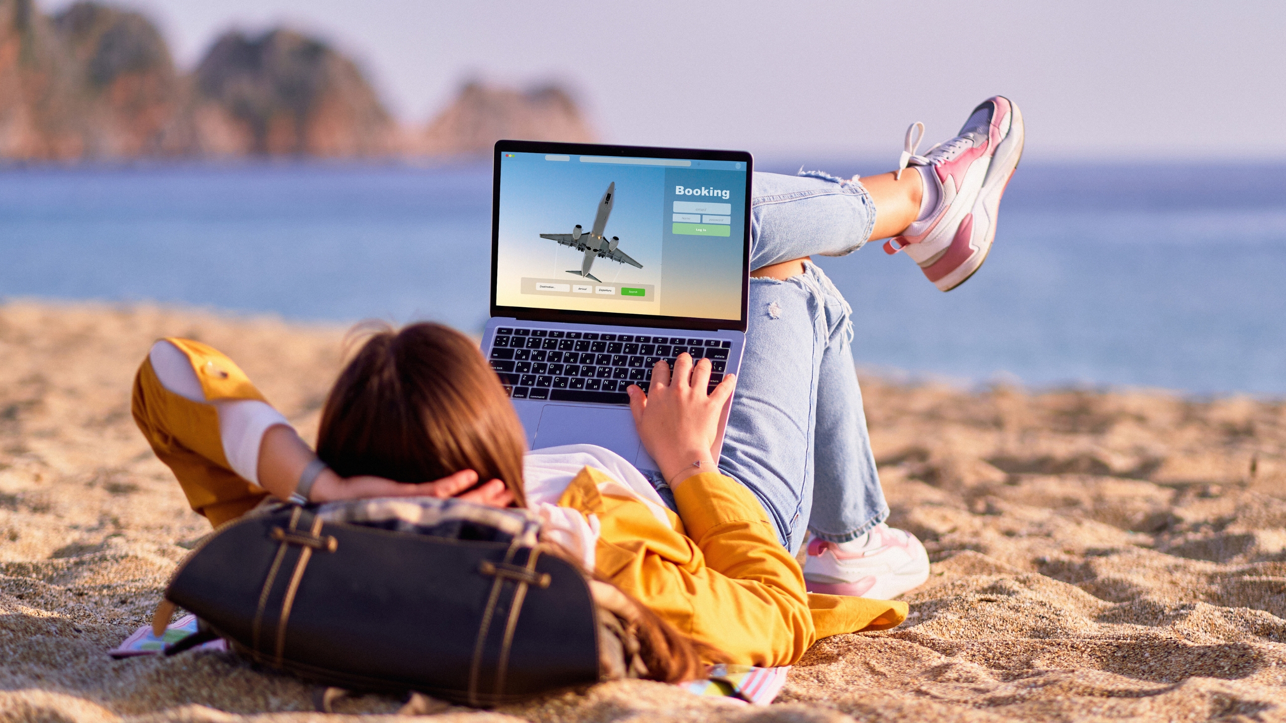 A person lying on a sandy beach, using a laptop with a Sustainable Business Audit website on the screen. The ocean and rocks are visible in the background. The person is relaxed, wearing casual clothes and sneakers.