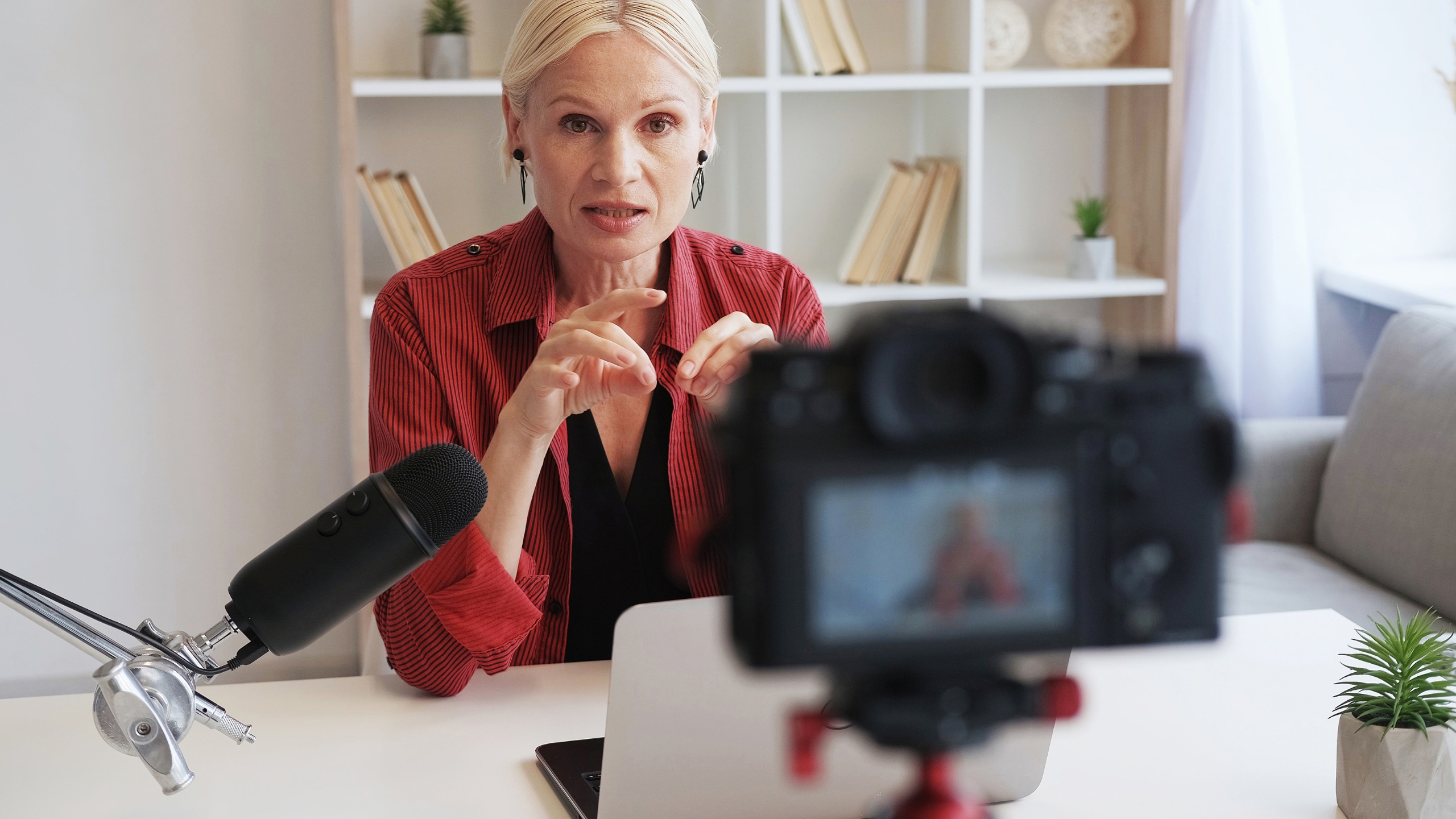 A woman with blonde hair sits at a desk with a microphone and laptop, sharing tips on how to nail your marketing while speaking to a camera in a bright, modern room with shelves and books in the background.