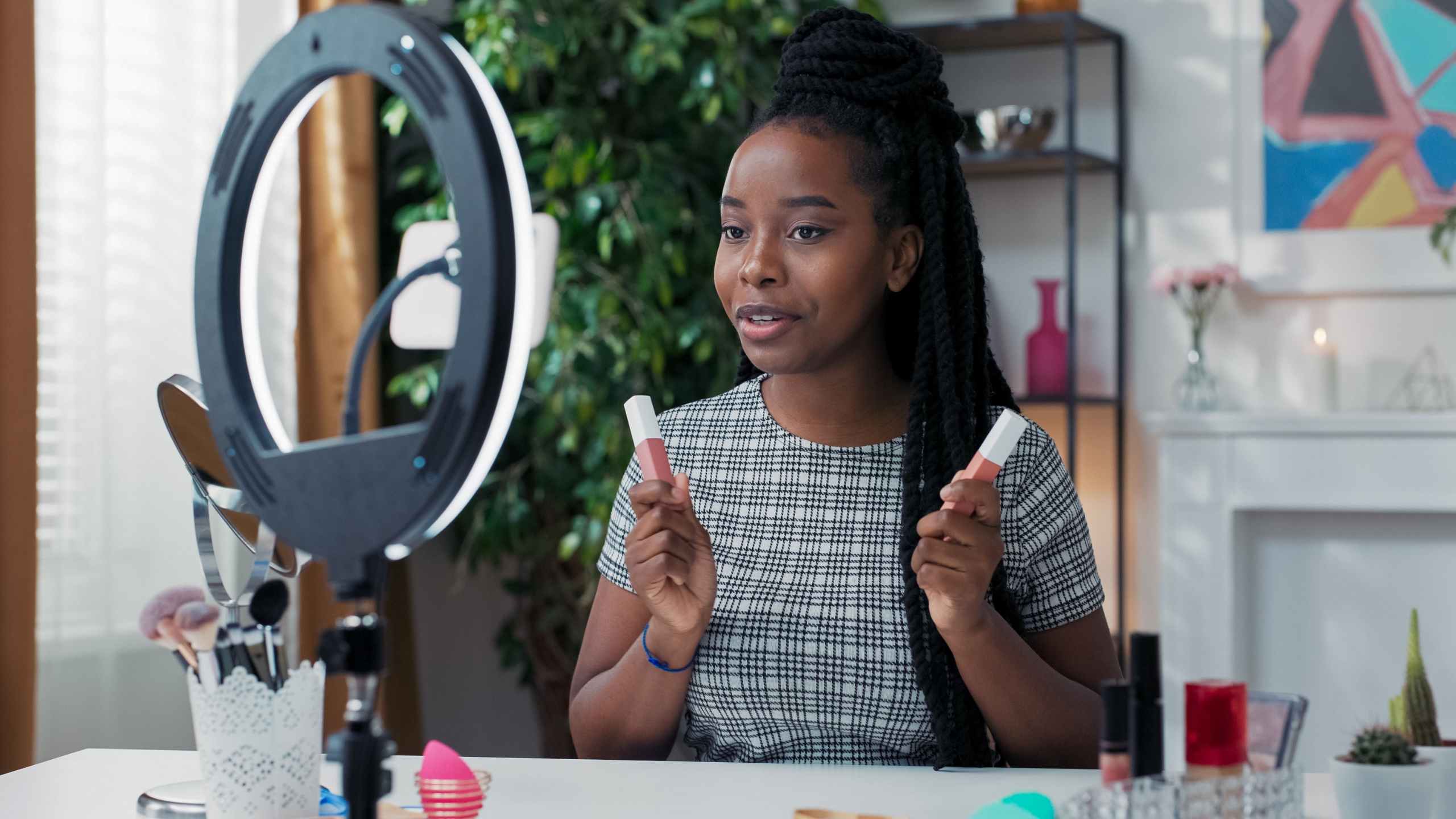 A woman sits at a desk with makeup products, holding two lipsticks and speaking in front of a ring light and camera—likely sharing tips to help her followers learn how to boost their beauty skills or even make money online.