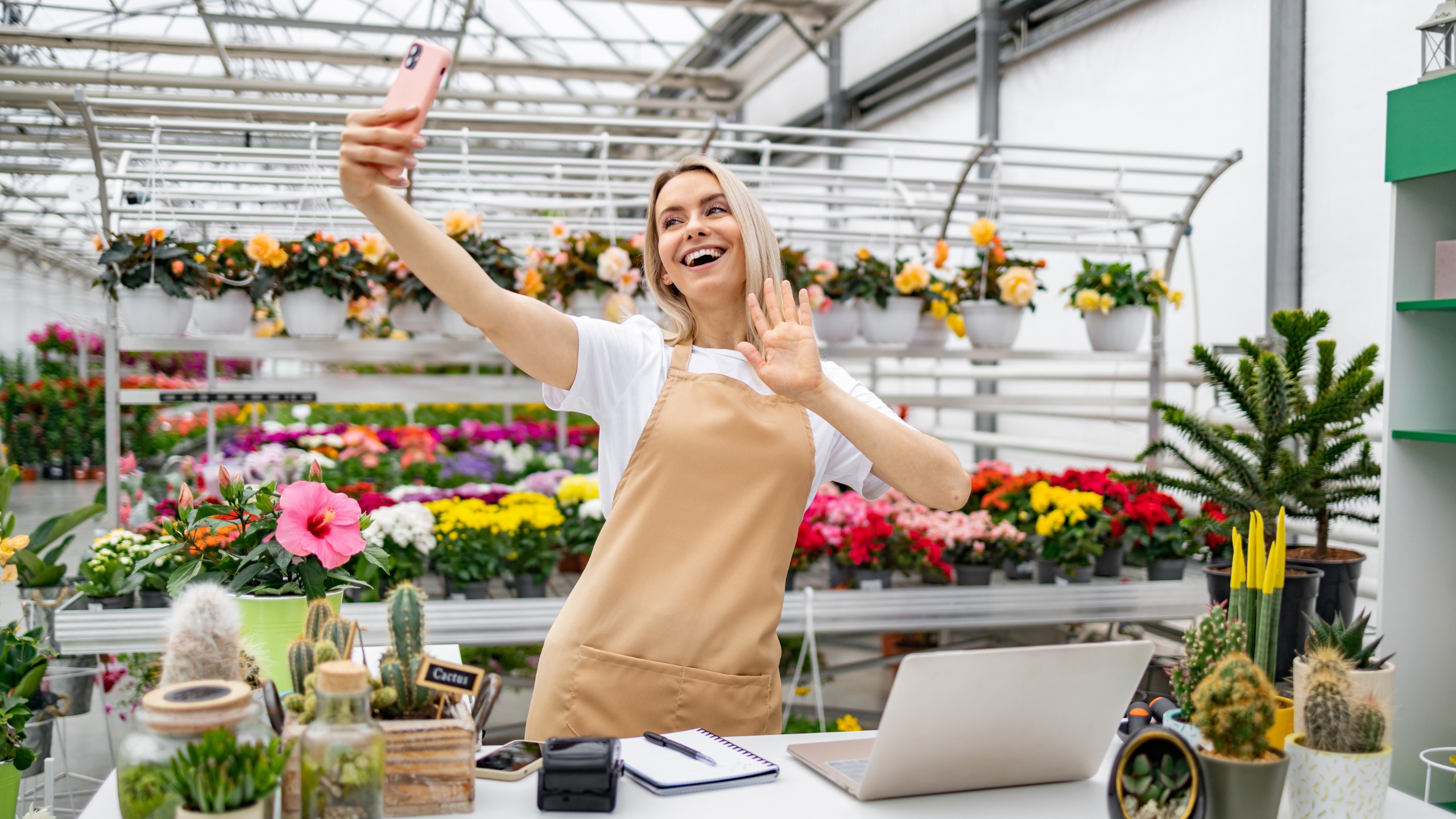A smiling woman in an apron takes a selfie and waves in her flower shop, surrounded by colorful plants and flowers. A laptop, notebook, and camera on the counter hint at her creative approach to building passive income.