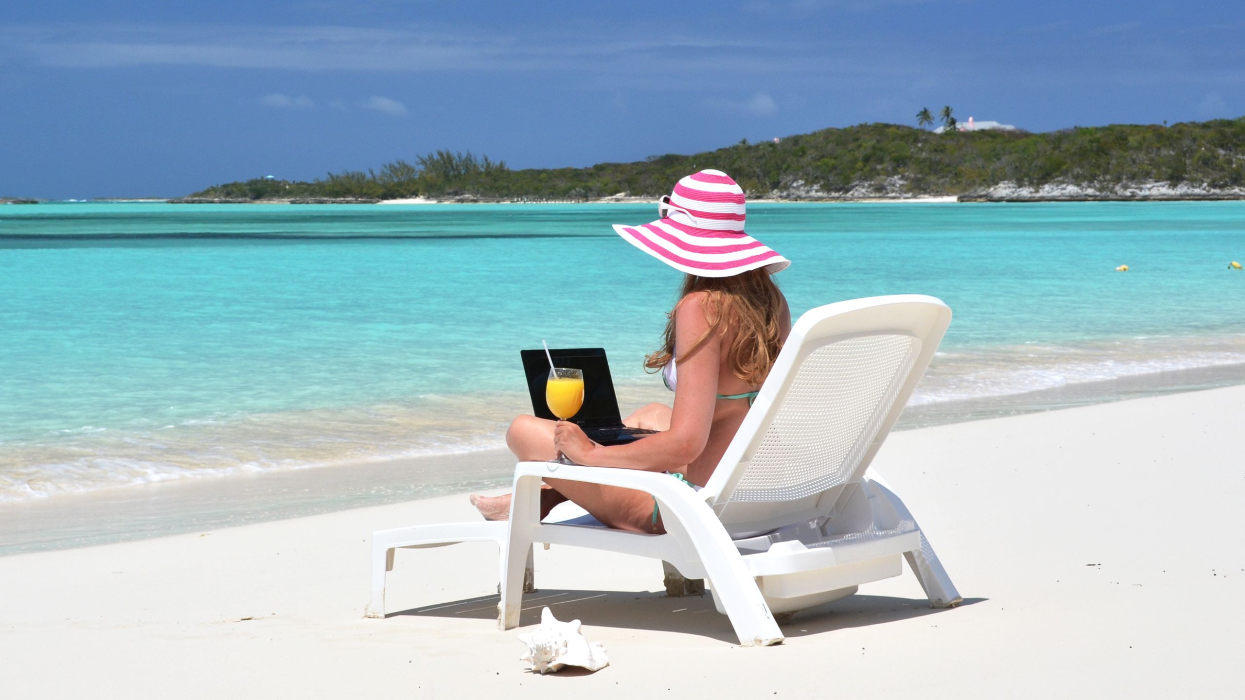 A woman in a pink-and-white striped sunhat sits on a lounge chair at a tropical beach, working on her laptop and holding a drink, exploring AI for business as turquoise water and green islands create the perfect backdrop.