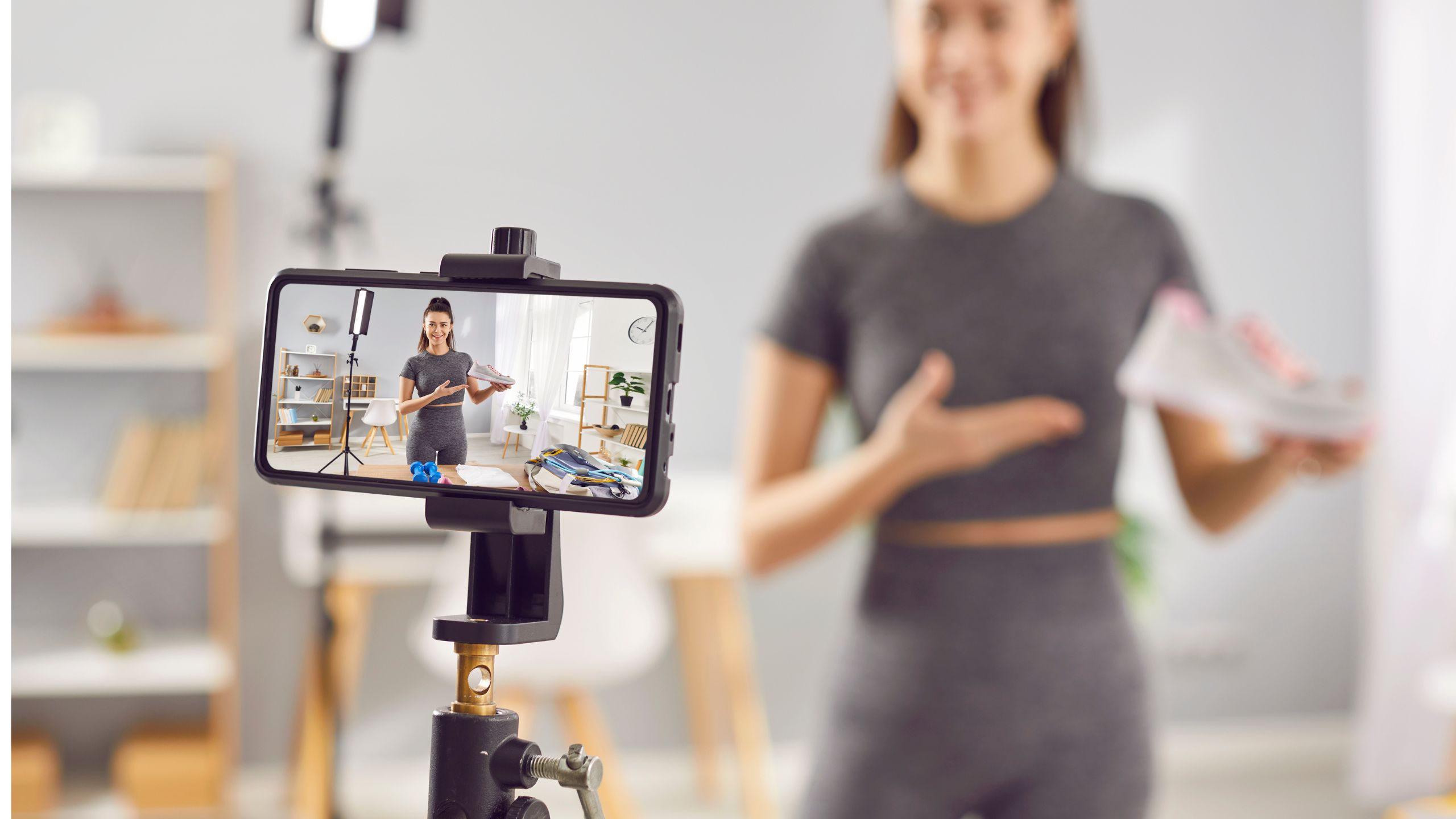 A woman in athletic wear stands in a room, holding a sneaker and gesturing toward it while filming herself for her master podcast page. The phone screen shows her presenting the shoe in a bright, home setting.