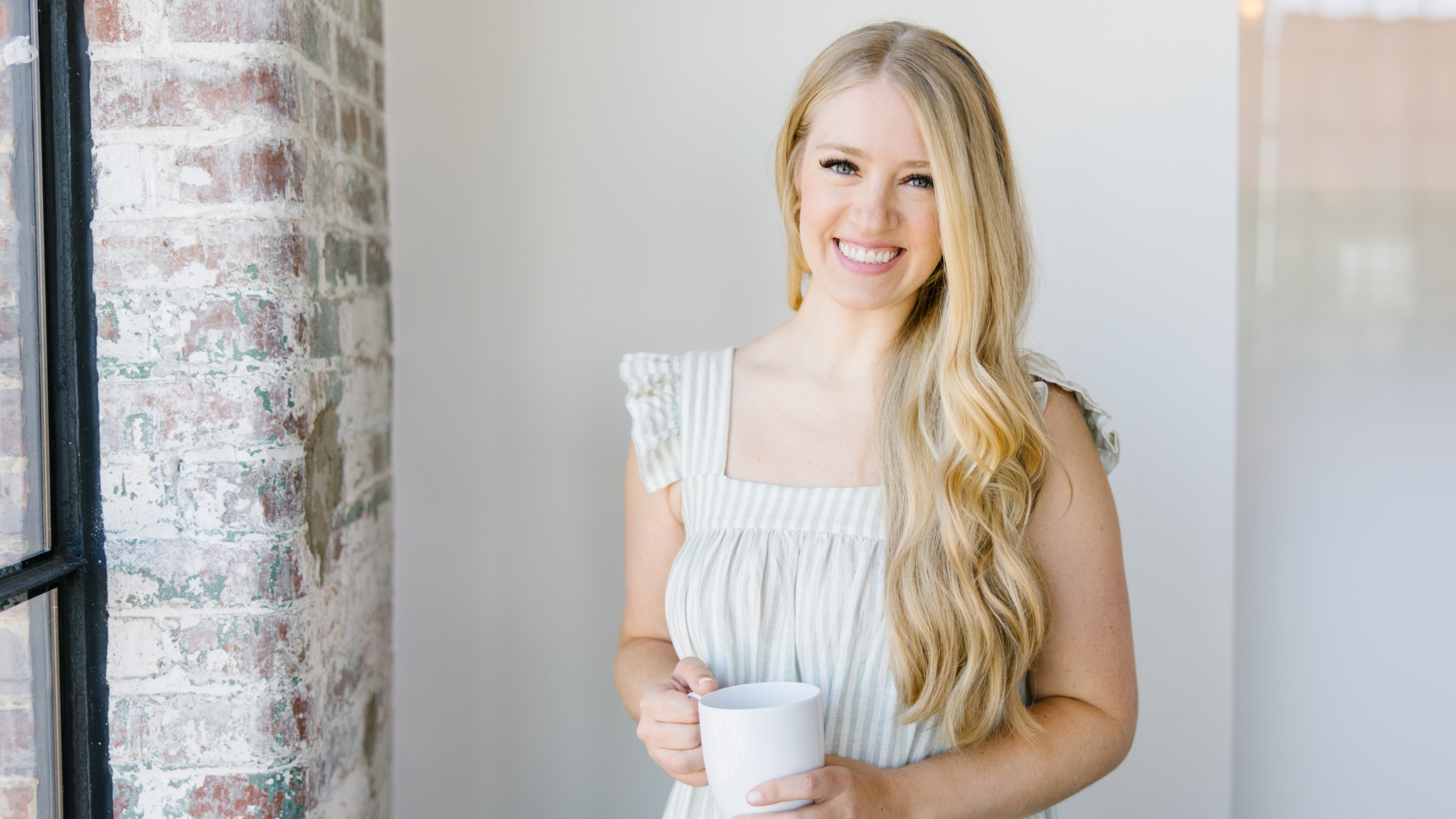 Stephanie May Willson, a young woman with long blonde hair, stands by a brick wall in a bright, airy room, wearing a light-colored sleeveless dress and holding a white mug as she smiles at the camera.