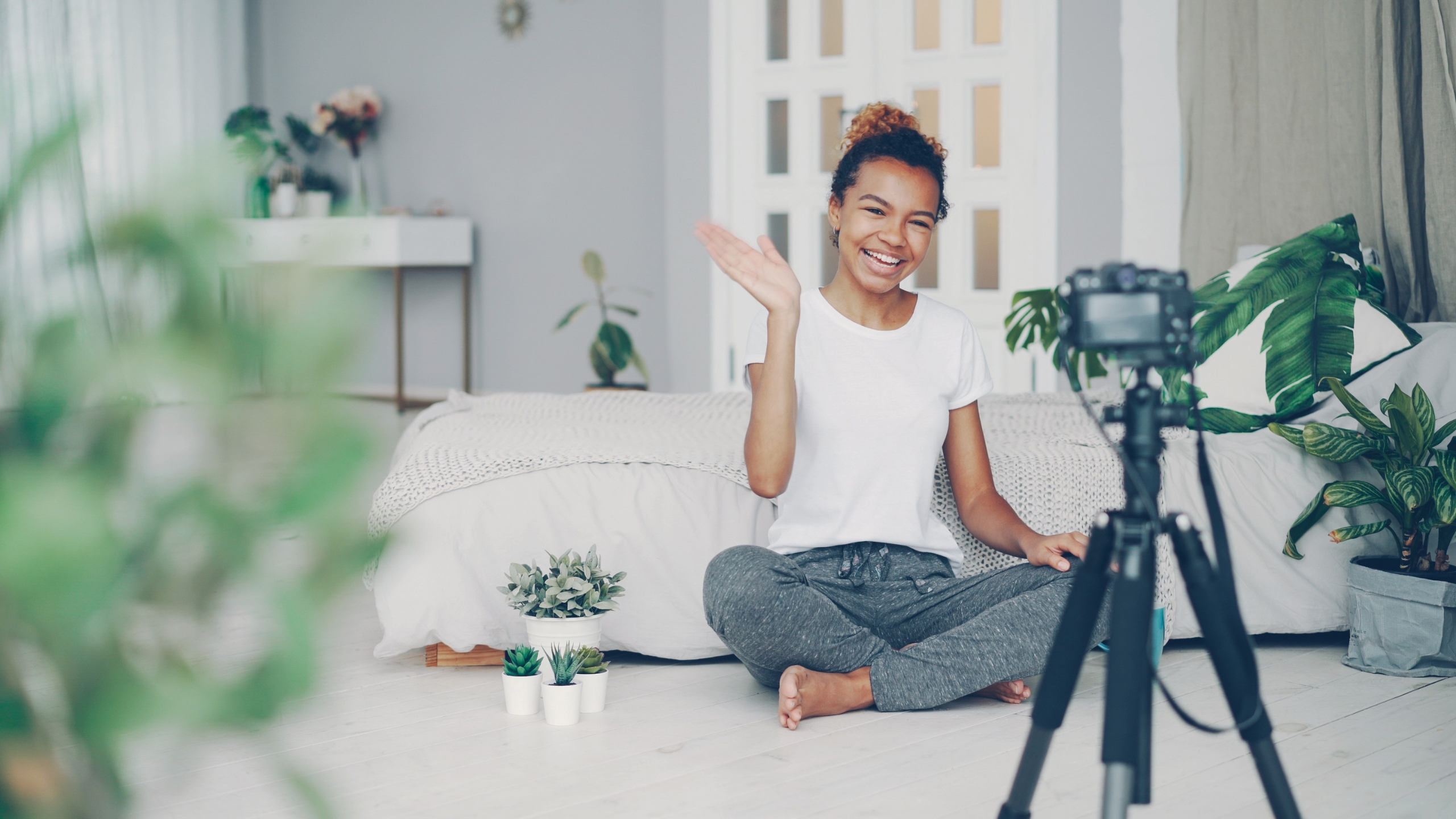 A smiling woman sits cross-legged on the floor in front of a bed, waving at a camera on a tripod. Perfect for course creators or coaches, she’s filming Meta ads in a bright, cozy room filled with plants and modern decor.