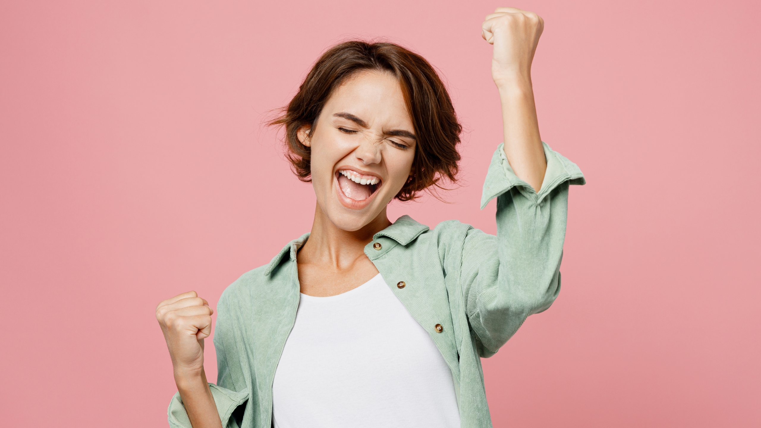 A young woman with short brown hair celebrates joyfully, eyes closed, smiling with mouth open and raising both fists in the air, standing against a pink background—capturing the thrill of a big sales win in marketing.