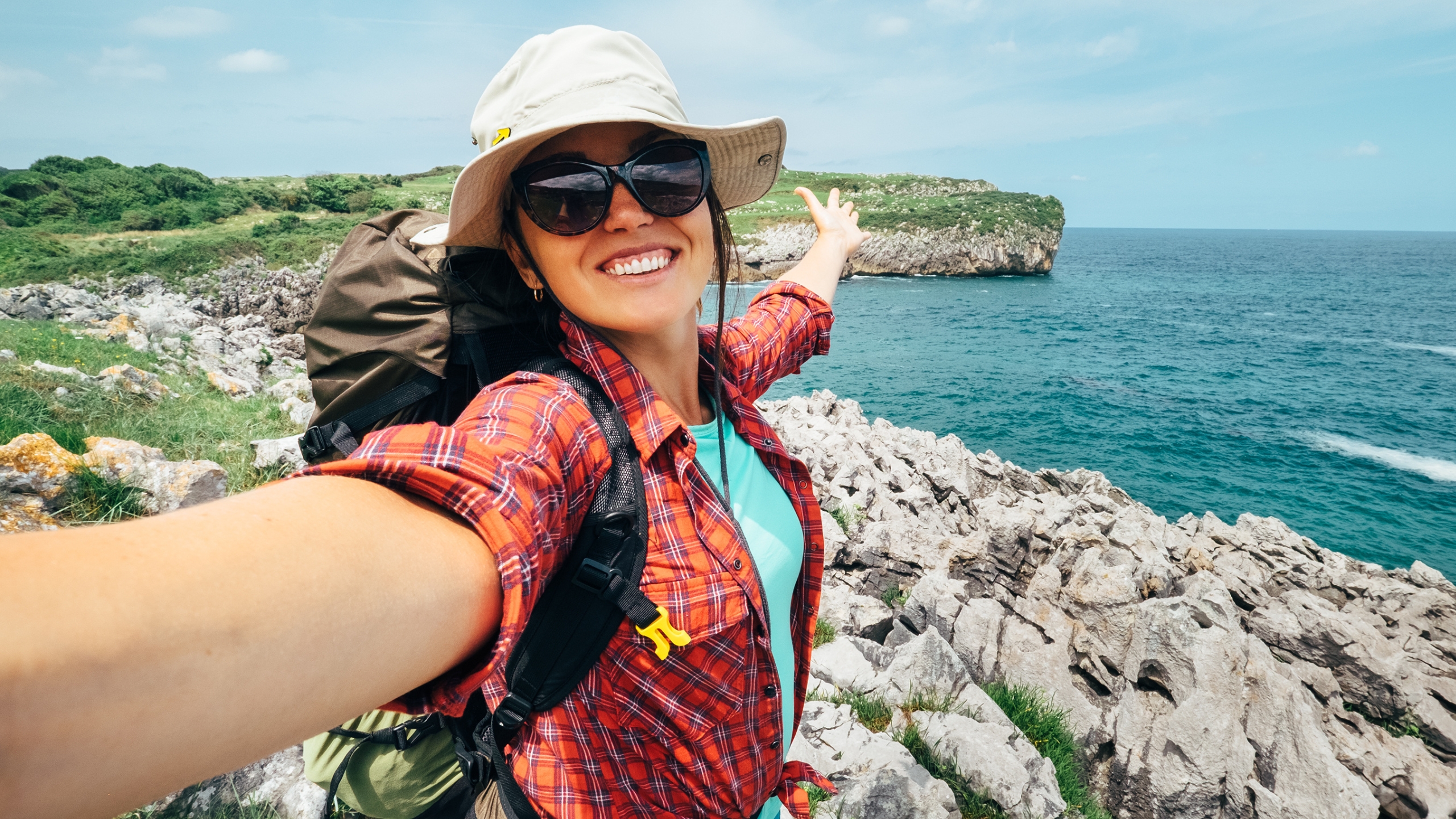A smiling woman in sunglasses, a hat, and a plaid shirt takes a selfie while hiking near the rocky coastline, dreaming of her consistent $10K months as she points toward the sea with confidence and adventure in her eyes.