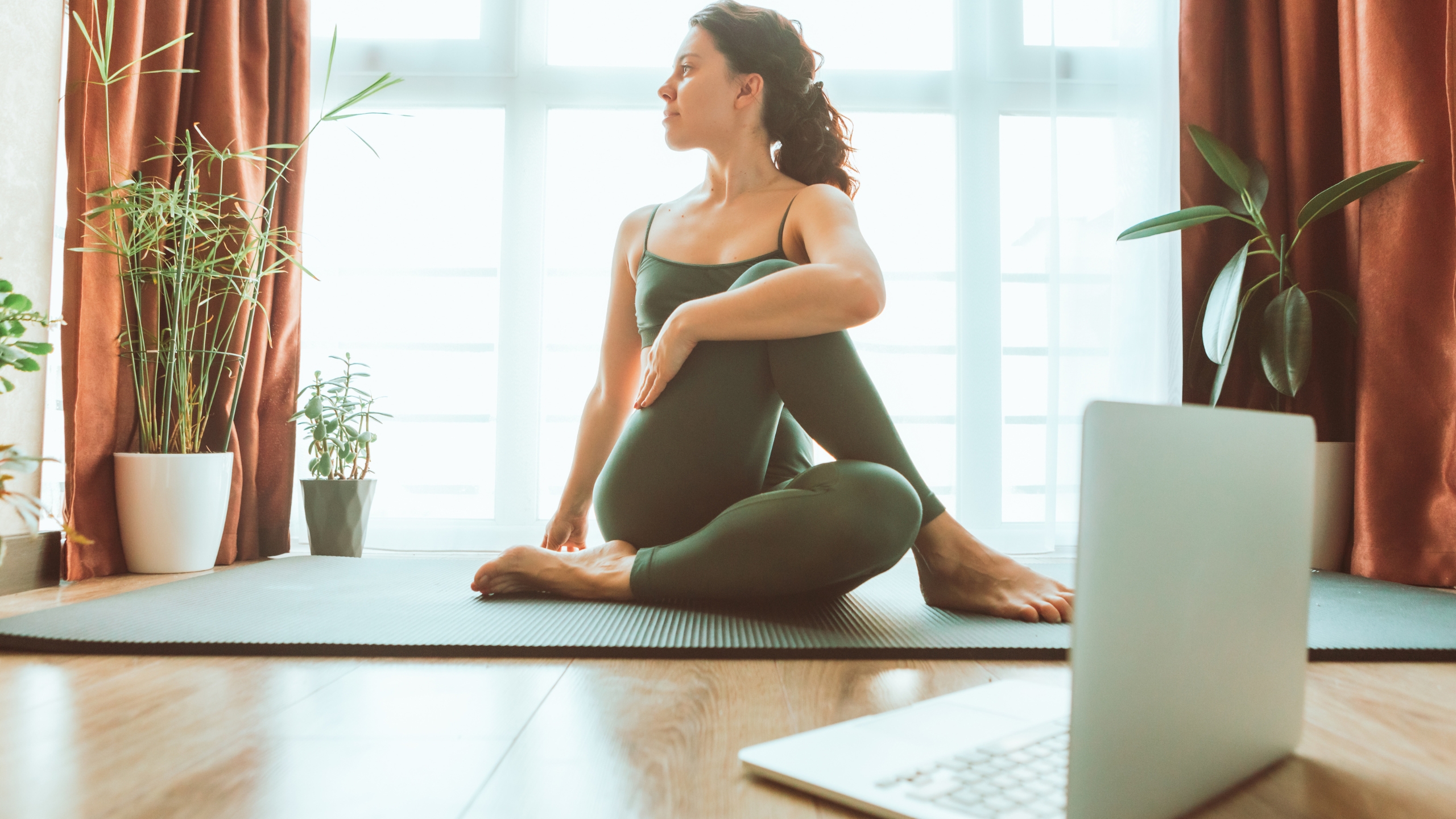 A woman in athletic wear practices a seated yoga twist on a mat indoors, surrounded by houseplants and sunlight, while following behind-the-scenes launch strategies on her laptop.