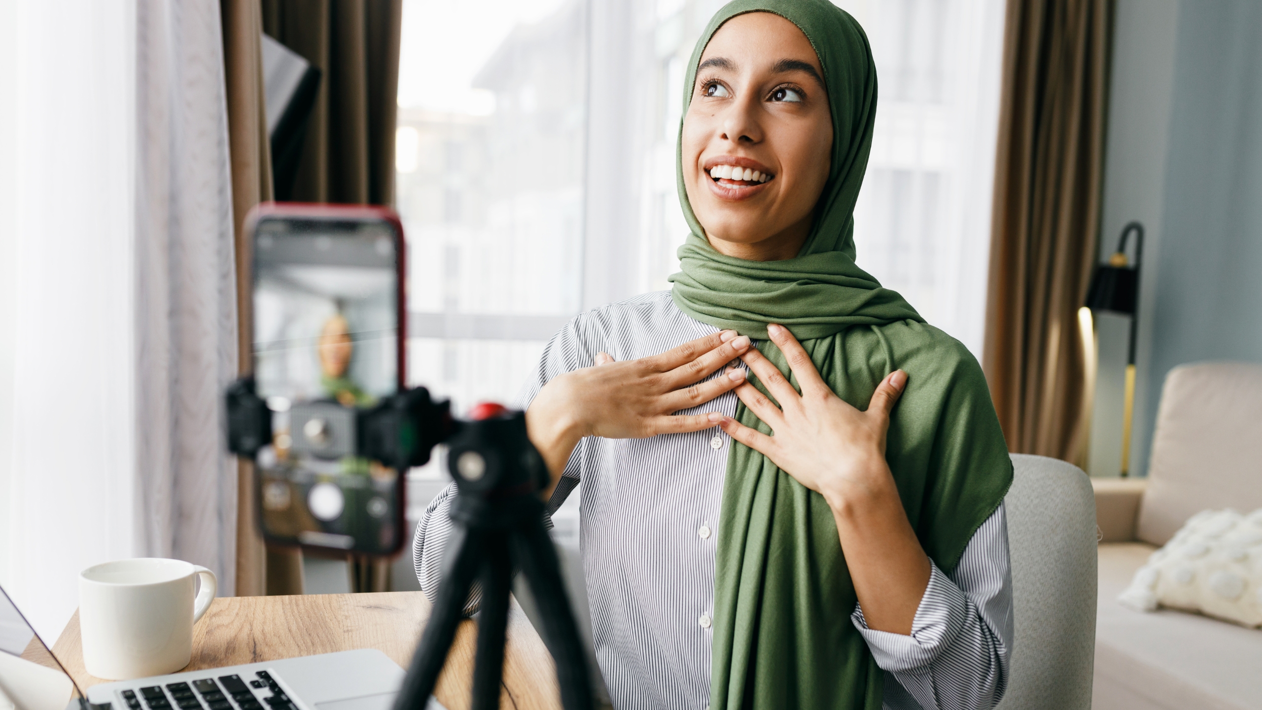 A woman in a green hijab sits at a desk, smiling and gesturing while recording a video on her smartphone, which is mounted on a tripod. As she shares her thoughts on decision making and personal growth, a laptop and a coffee cup sit on the table in front of her.