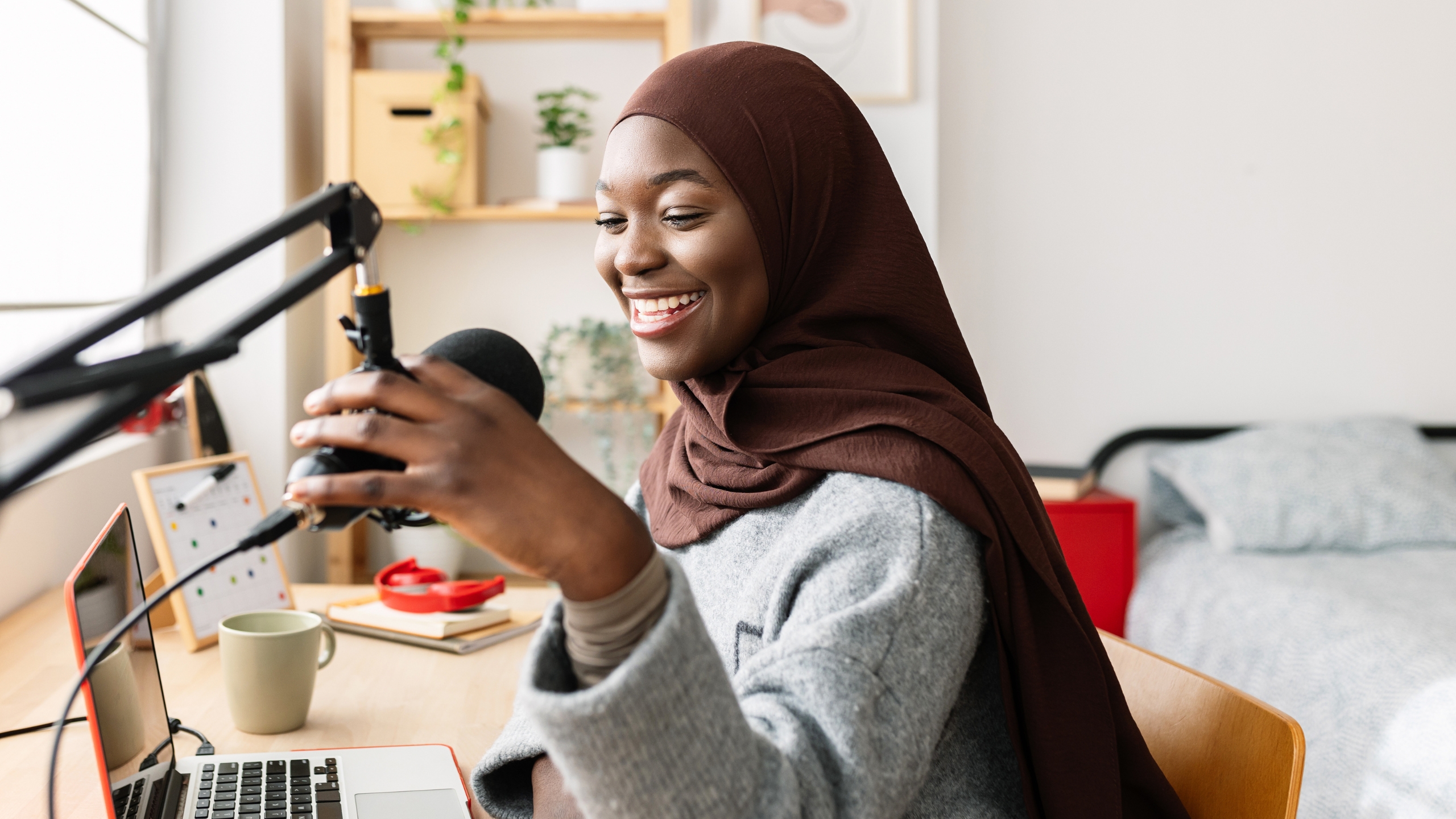 A woman wearing a brown hijab smiles while speaking into a microphone at her desk, with a laptop, notebook, and coffee mug beside her, in a bright, cozy room—perfectly capturing the positive energy behind increasing conversions and engaging views.