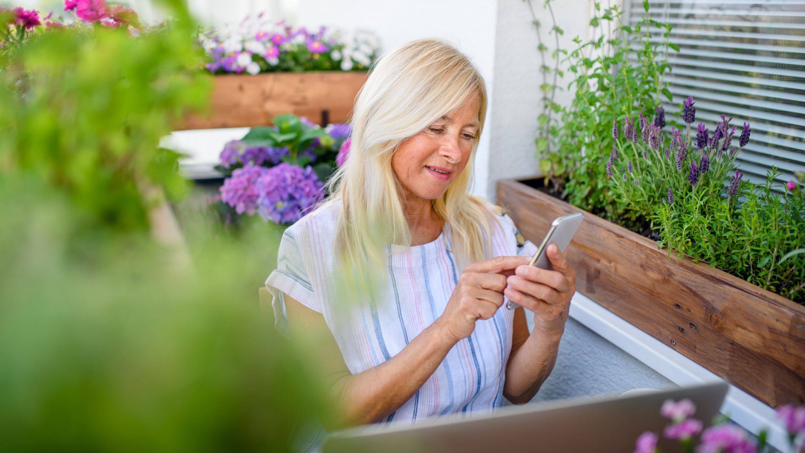 Older woman with blonde hair sits outdoors among potted flowers, smiling and using a smartphone—perhaps exploring Instagram Trial Reels to build your audience. A laptop is visible in front of her, and the setting appears to be a balcony or patio garden.