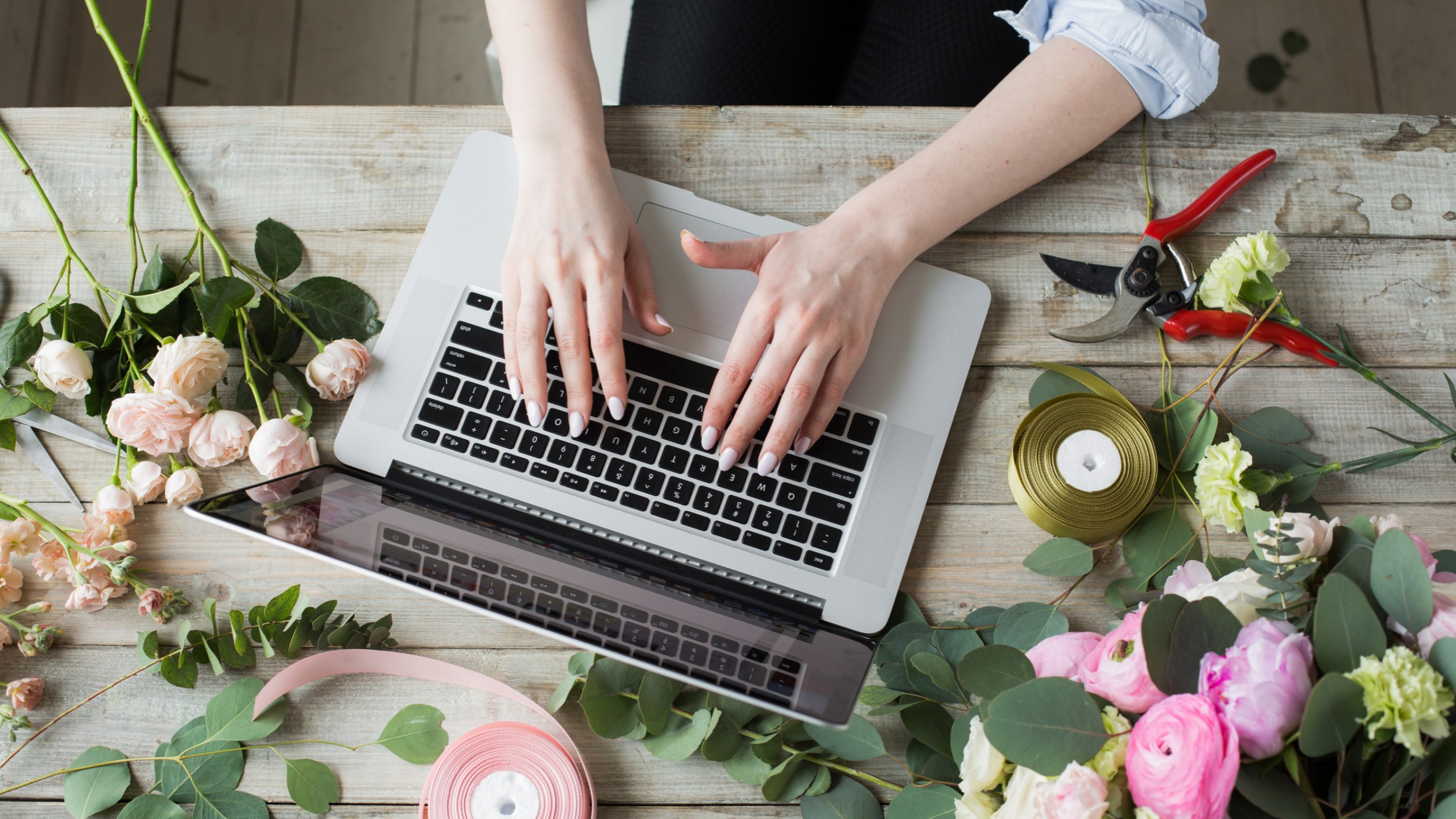 A person typing on a laptop surrounded by flowers, floral shears, and ribbon on a wooden table while working on an online course launch.