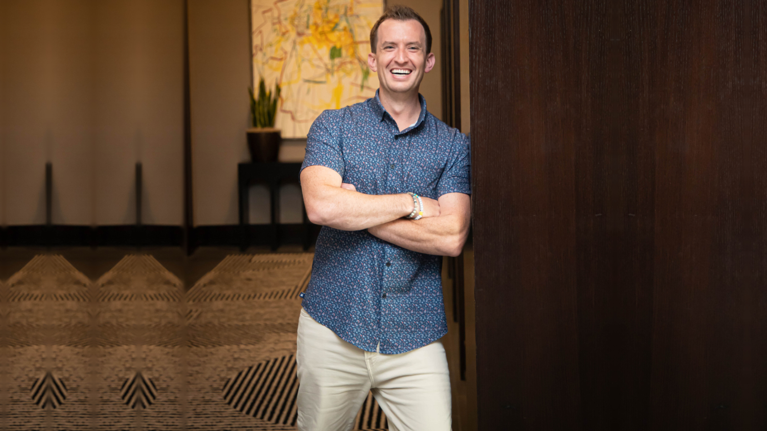 Brad Bizjack stands with his arms crossed and a smile, leaning against a dark wooden wall while wearing a blue patterned shirt and tan pants to represent entrepreneur fatigue.